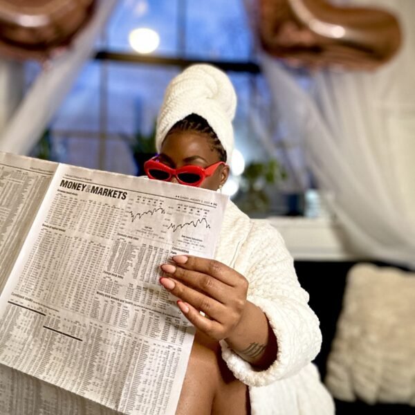 LAUNA in a robe and hair towel wearing red cat-eye sunglasses, reading the Money and Markets pages