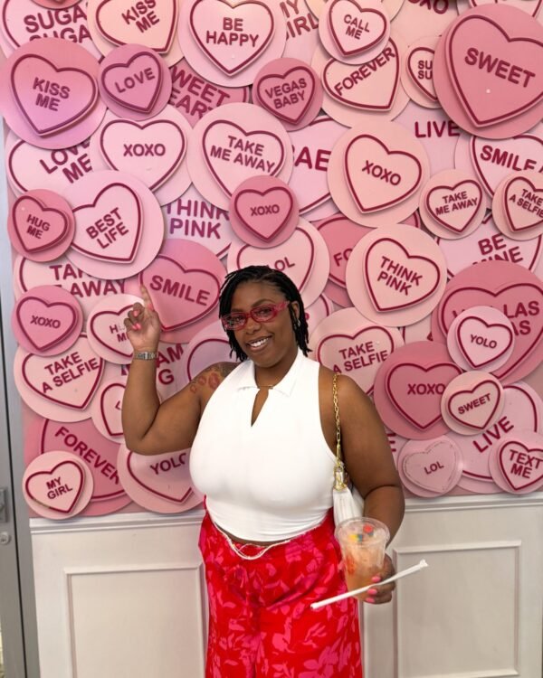 Launa in a white top and red floral skirt in front of a pink candy-heart wall