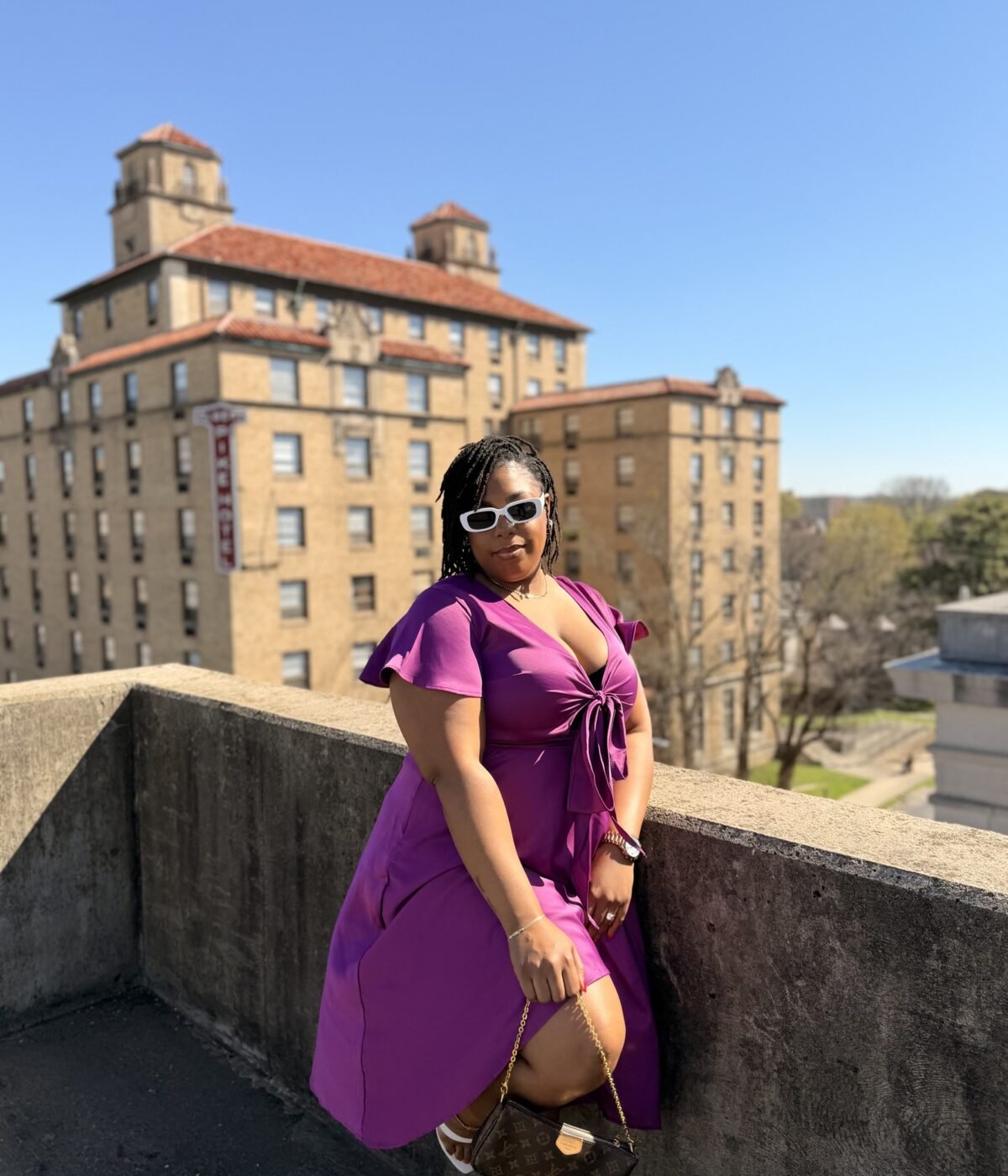 LAUNA in a magenta wrap dress and sunglasses on a rooftop, city building behind her