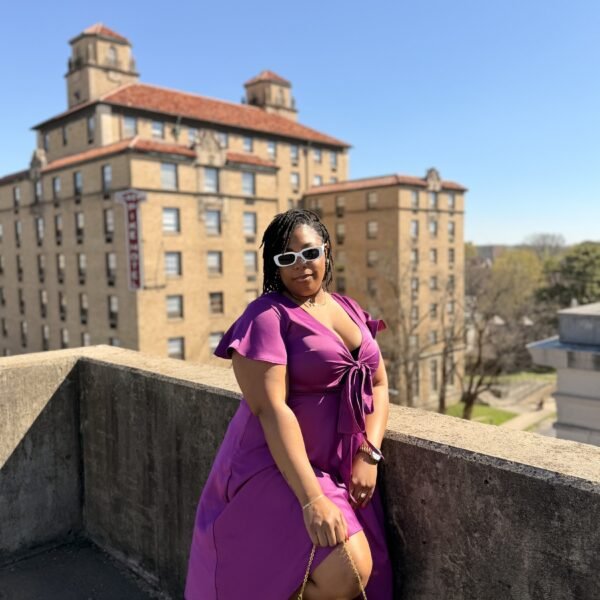 LAUNA in a magenta wrap dress and sunglasses on a rooftop, city building behind her