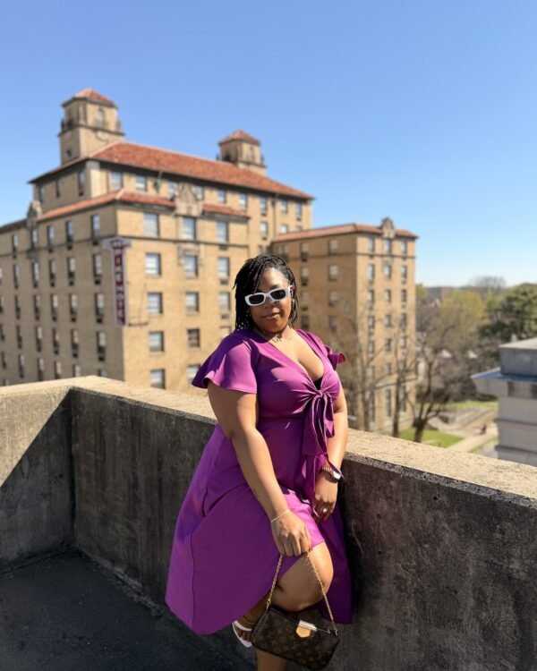 LAUNA in a magenta wrap dress and sunglasses on a rooftop, city building behind her