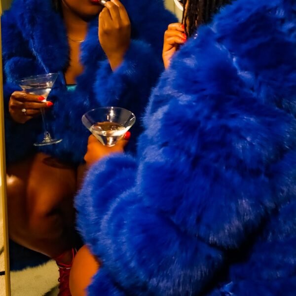 Launa in a cobalt faux-fur coat with a cocktail, reflected in a mirror