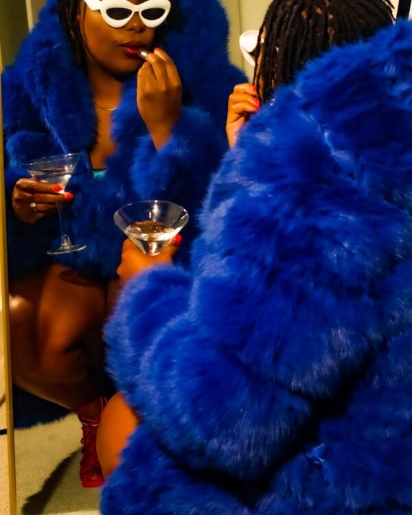 Launa in a cobalt faux-fur coat with a cocktail, reflected in a mirror