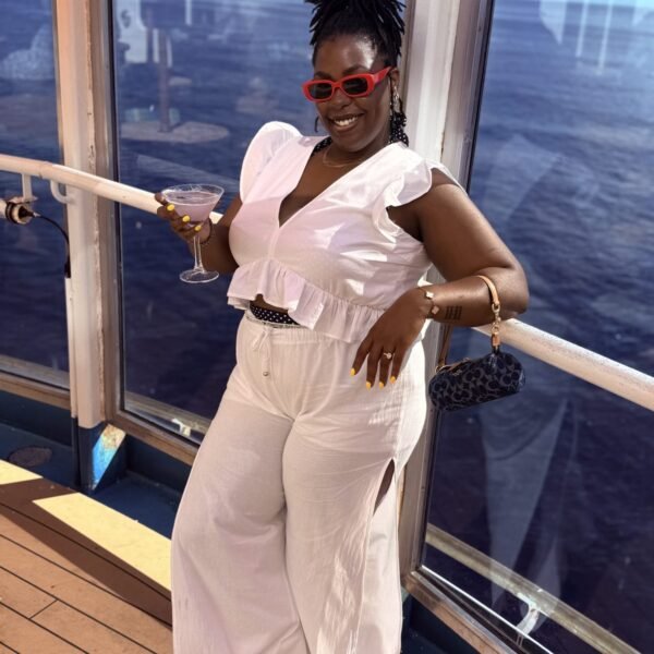 Launa in a white jumpsuit and red sunglasses by a window over open water